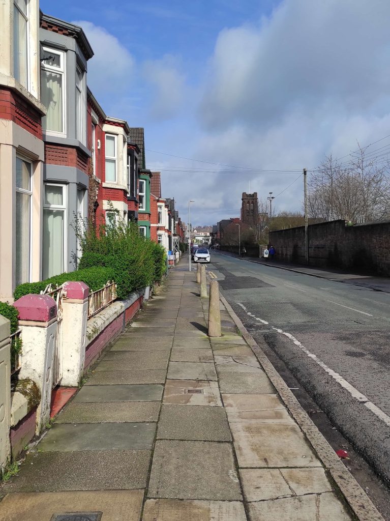 Liverpool et les Beatles : musées, visites, et lieux emblématiques à ne pas manquer 7 La rue de Penny Lane avec maisons à bow window sur la gauche, la route goudronnée sur la droite et une cathédrale au fond.