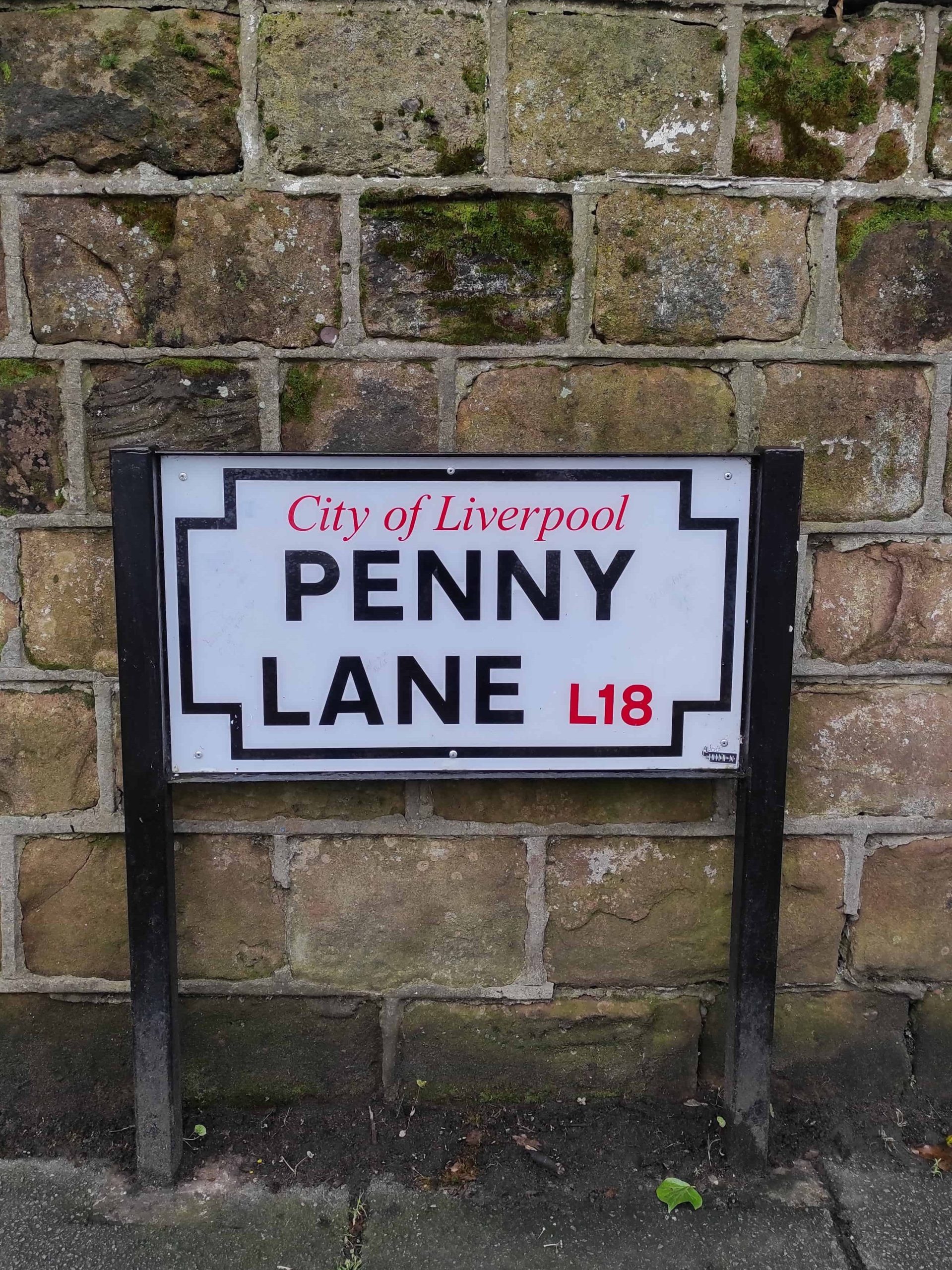 Liverpool et les Beatles : musées, visites, et lieux emblématiques à ne pas manquer 6 Panneau de la rue de Penny Lane blanc avec écriture noire, devant mur en pierre