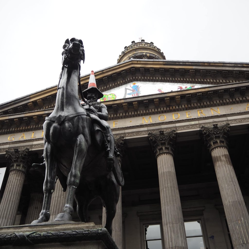 3 circuits parfaits pour visiter Glasgow le temps d'un long weekend 16 Vue en contre plongée d'une statue d'un homme sur un cheval devant un bâtiment en pierre type panthéon avec des colonnes grecques. Le bâtiment porte un nom en lettres dorées "GALLERY OF MODERN ART" et la statue porte un plot de chantier orange et blanc sur la tête.