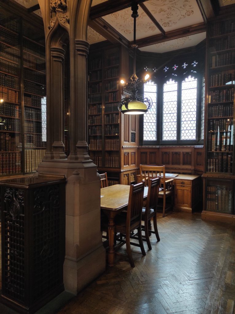 4 musées à Manchester à découvrir absolument sans débourser un centime 17 Salle de travail dans la bibliothèque John Rylands avec chaise et table en bois, grands vitraux, colonnes en pierre et livres dans grandes bibliothèques