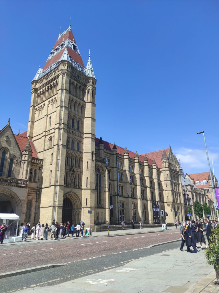 4 musées à Manchester à découvrir absolument sans débourser un centime 10 Extérieur du Manchester Museum. Grand bâtiment en pierres beiges avec toit rouge et détails dentelés sur le toit d'une tour. Rue au premier plan avec passants sur les trottoirs