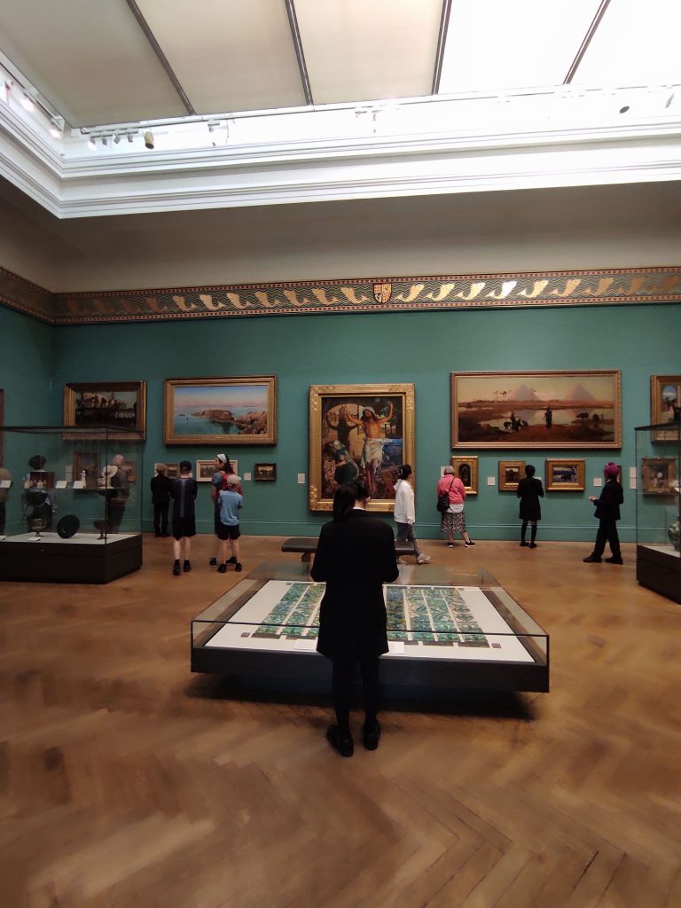4 musées à Manchester à découvrir absolument sans débourser un centime 9 Grande pièce avec très grandes peintures exposées. Au centre de la pièce sur parquet en bois, un présentoir avec une oeuvre. Quelques personnes admirent les peintures.